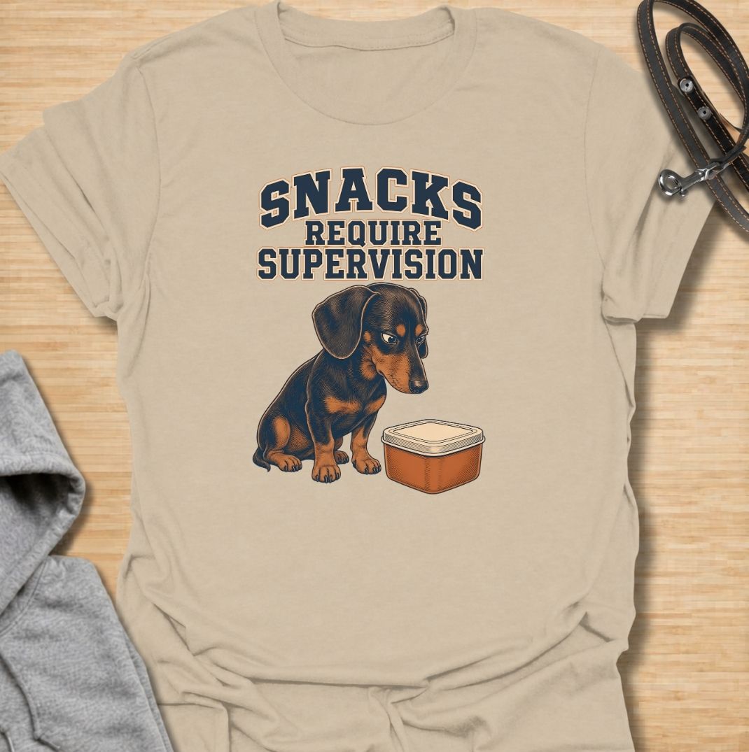 A funny dachshund T-shirt for anyone whose dog believes every snack requires close supervision. Featuring the phrase “Snacks Require Supervision” with a watchful dachshund beside a treat container, this design captures the breed’s famously food-focused personality. Perfect for dachshund moms, dachshund dads, sausage dog owners, and anyone who can’t open a snack without attracting immediate attention. A playful everyday tee for fans of dachshund humor, wiener dogs, and permanently hungry little companions.
