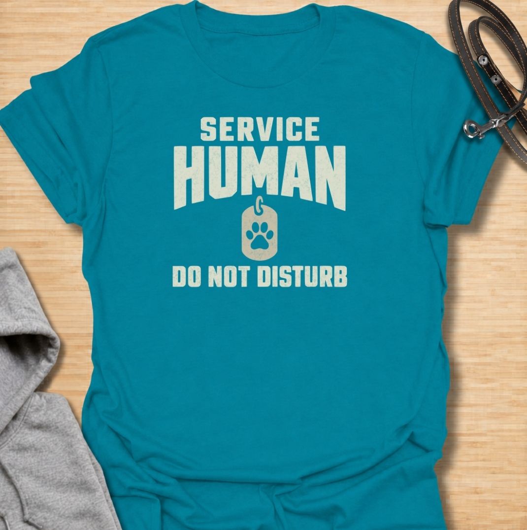 A funny dog T-shirt for anyone who knows their main role at home is serving the dog. Featuring the phrase “Service Human — Do Not Disturb” with a simple paw-print tag graphic, this clean design turns the service-dog idea on its head with dry pet-owner humor. Perfect for dog moms, dog dads, rescue dog owners, and anyone whose four-legged boss expects constant attention. A playful everyday tee for fans of funny dog sayings, pet parent humor, and general dog lover gifts.