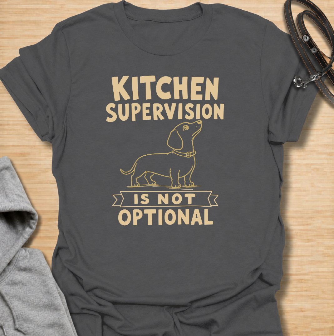 A funny dachshund T-shirt for anyone who never enters the kitchen without their four-legged supervisor close behind. Featuring a clean vintage-inspired dachshund illustration and the phrase “Kitchen Supervision Is Not Optional,” this design captures one of the breed’s most familiar habits. Perfect for dachshund moms, dachshund dads, sausage dog owners, and anyone whose dog appears the moment food is involved. A playful everyday tee for fans of dachshund humor, wiener dogs, and food-obsessed pups.