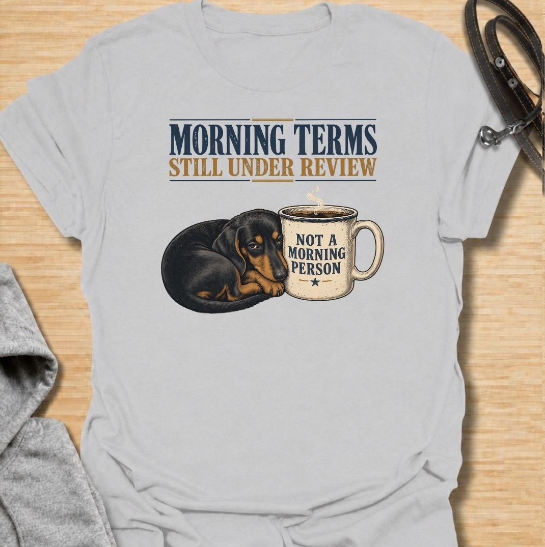 A funny dachshund T-shirt for anyone whose mornings require coffee, patience, and very limited conversation. Featuring a sleepy dachshund curled beside a coffee mug with the phrases “Morning Terms Still Under Review” and “Not a Morning Person,” this design has a dry, relatable sense of humor. Perfect for dachshund moms, dachshund dads, coffee lovers, and anyone who prefers a slow start to the day. A fun everyday tee combining dachshund humor, coffee culture, and an unmistakably grumpy morning mood.