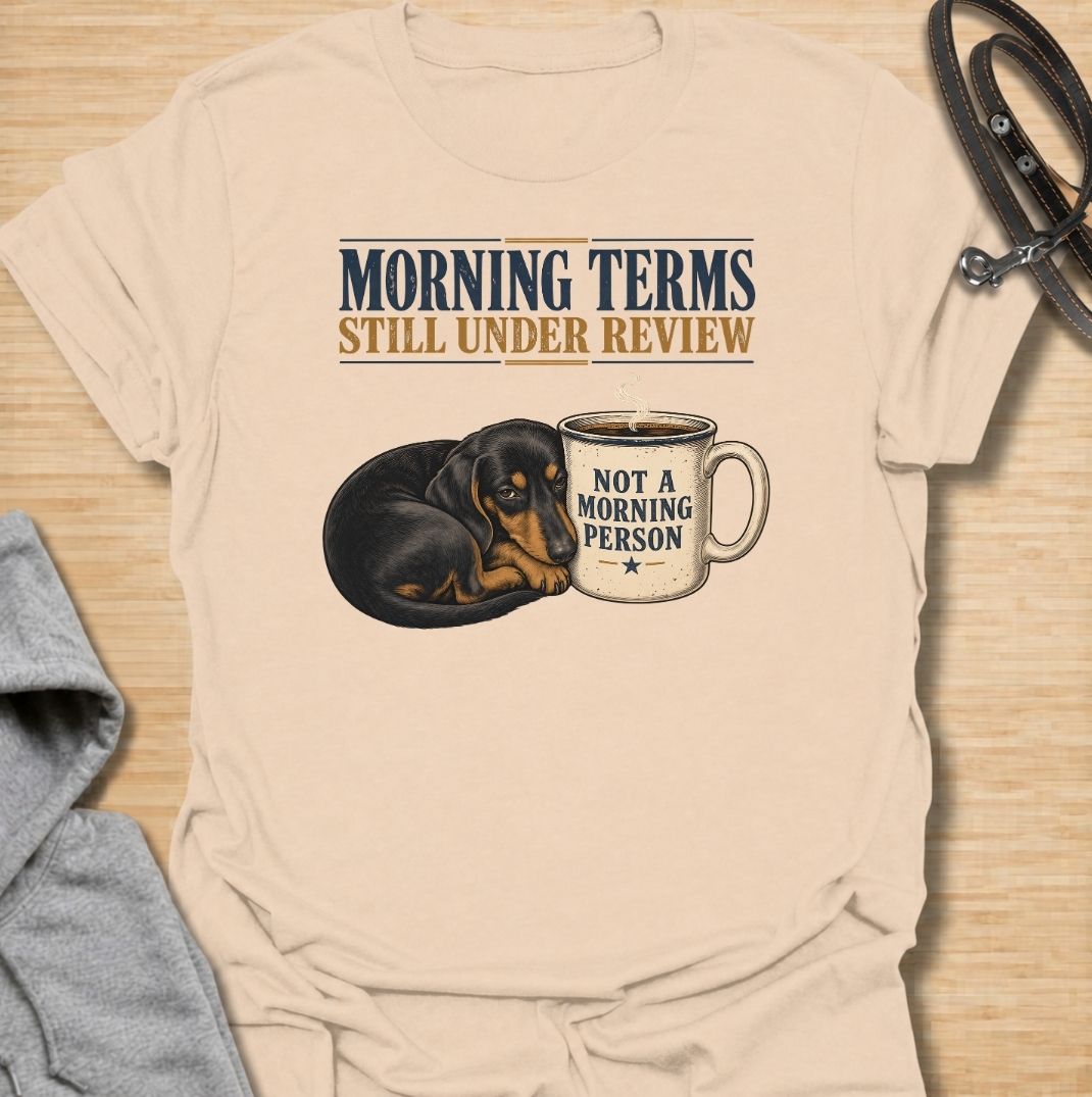A funny dachshund T-shirt for anyone whose mornings require coffee, patience, and very limited conversation. Featuring a sleepy dachshund curled beside a coffee mug with the phrases “Morning Terms Still Under Review” and “Not a Morning Person,” this design has a dry, relatable sense of humor. Perfect for dachshund moms, dachshund dads, coffee lovers, and anyone who prefers a slow start to the day. A fun everyday tee combining dachshund humor, coffee culture, and an unmistakably grumpy morning mood.