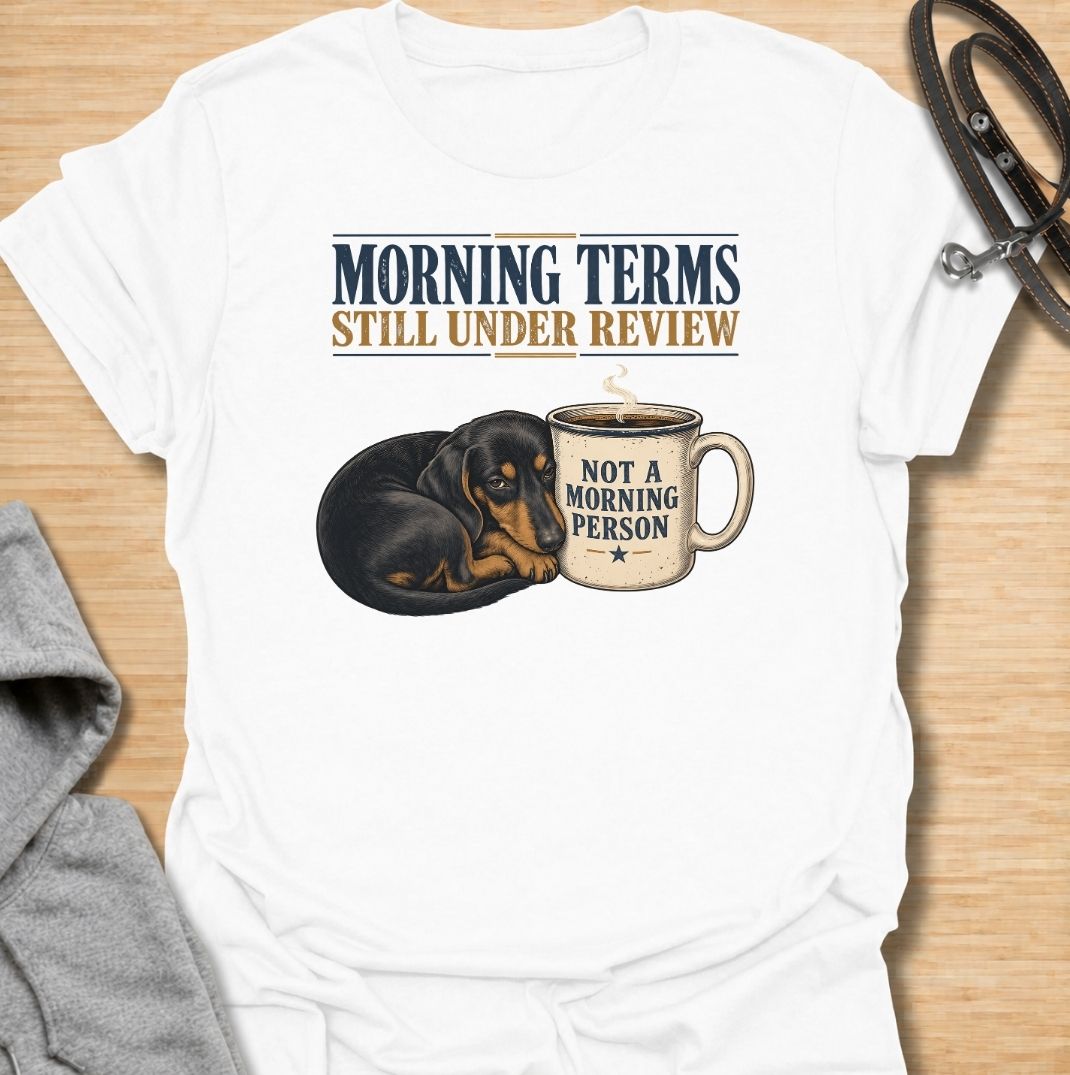 A funny dachshund T-shirt for anyone whose mornings require coffee, patience, and very limited conversation. Featuring a sleepy dachshund curled beside a coffee mug with the phrases “Morning Terms Still Under Review” and “Not a Morning Person,” this design has a dry, relatable sense of humor. Perfect for dachshund moms, dachshund dads, coffee lovers, and anyone who prefers a slow start to the day. A fun everyday tee combining dachshund humor, coffee culture, and an unmistakably grumpy morning mood.