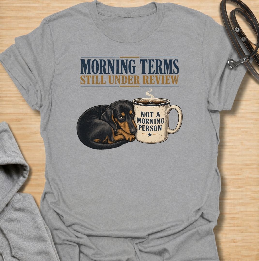 A funny dachshund T-shirt for anyone whose mornings require coffee, patience, and very limited conversation. Featuring a sleepy dachshund curled beside a coffee mug with the phrases “Morning Terms Still Under Review” and “Not a Morning Person,” this design has a dry, relatable sense of humor. Perfect for dachshund moms, dachshund dads, coffee lovers, and anyone who prefers a slow start to the day. A fun everyday tee combining dachshund humor, coffee culture, and an unmistakably grumpy morning mood.