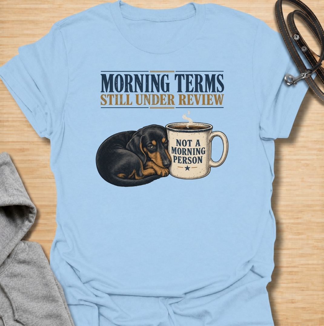 A funny dachshund T-shirt for anyone whose mornings require coffee, patience, and very limited conversation. Featuring a sleepy dachshund curled beside a coffee mug with the phrases “Morning Terms Still Under Review” and “Not a Morning Person,” this design has a dry, relatable sense of humor. Perfect for dachshund moms, dachshund dads, coffee lovers, and anyone who prefers a slow start to the day. A fun everyday tee combining dachshund humor, coffee culture, and an unmistakably grumpy morning mood.