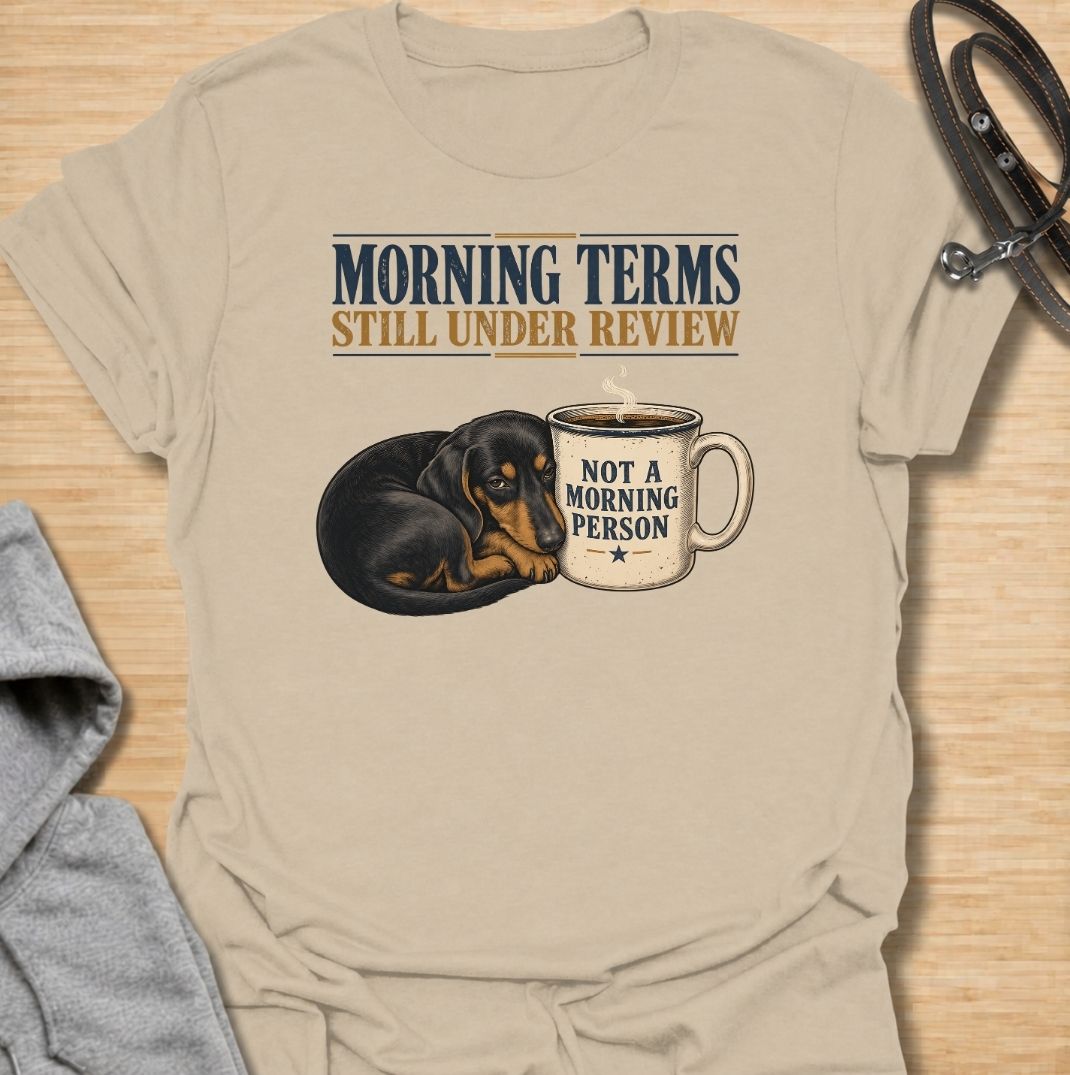 A funny dachshund T-shirt for anyone whose mornings require coffee, patience, and very limited conversation. Featuring a sleepy dachshund curled beside a coffee mug with the phrases “Morning Terms Still Under Review” and “Not a Morning Person,” this design has a dry, relatable sense of humor. Perfect for dachshund moms, dachshund dads, coffee lovers, and anyone who prefers a slow start to the day. A fun everyday tee combining dachshund humor, coffee culture, and an unmistakably grumpy morning mood.