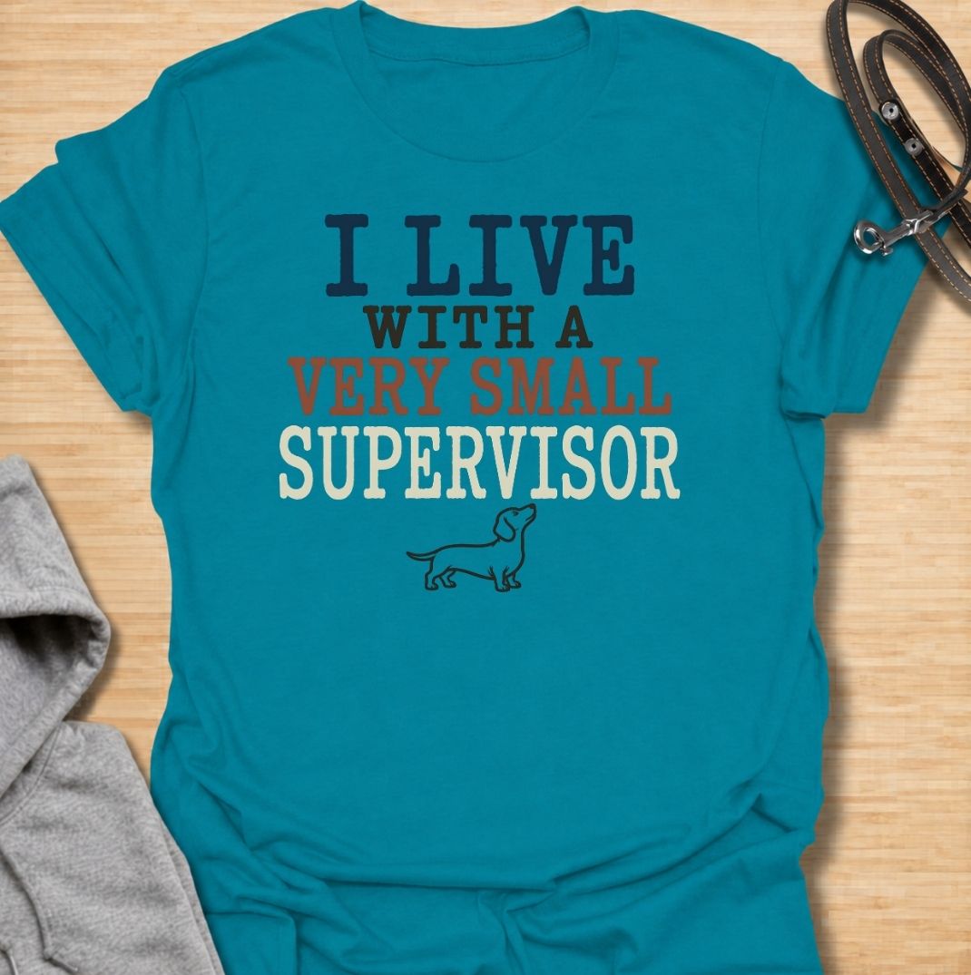 A funny dachshund T-shirt for anyone who lives with a very small dog who insists on overseeing absolutely everything. Featuring the phrase “I Live With a Very Small Supervisor” and a simple dachshund illustration, this design captures the breed’s curious, watchful personality perfectly. Ideal for dachshund moms, dachshund dads, sausage dog owners, and anyone whose little dog follows them from room to room. A playful everyday tee for fans of dachshund humor, wiener dogs, and devoted four-legged supervisors.
