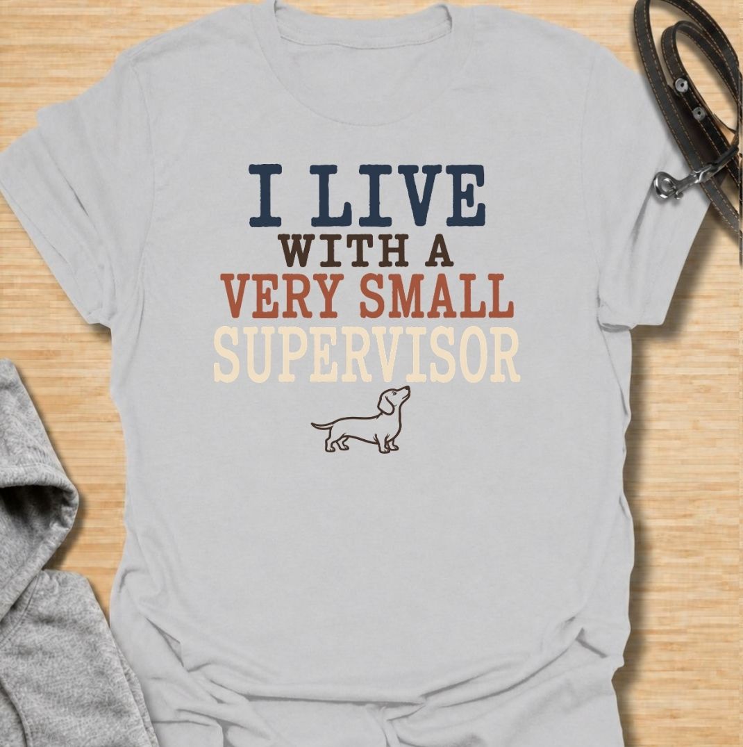 A funny dachshund T-shirt for anyone who lives with a very small dog who insists on overseeing absolutely everything. Featuring the phrase “I Live With a Very Small Supervisor” and a simple dachshund illustration, this design captures the breed’s curious, watchful personality perfectly. Ideal for dachshund moms, dachshund dads, sausage dog owners, and anyone whose little dog follows them from room to room. A playful everyday tee for fans of dachshund humor, wiener dogs, and devoted four-legged supervisors.