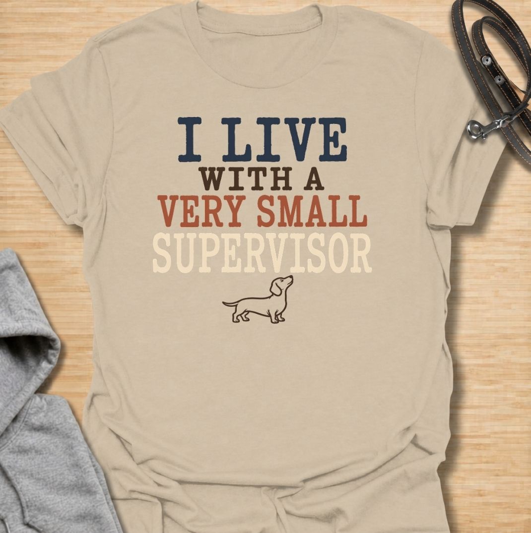 A funny dachshund T-shirt for anyone who lives with a very small dog who insists on overseeing absolutely everything. Featuring the phrase “I Live With a Very Small Supervisor” and a simple dachshund illustration, this design captures the breed’s curious, watchful personality perfectly. Ideal for dachshund moms, dachshund dads, sausage dog owners, and anyone whose little dog follows them from room to room. A playful everyday tee for fans of dachshund humor, wiener dogs, and devoted four-legged supervisors.