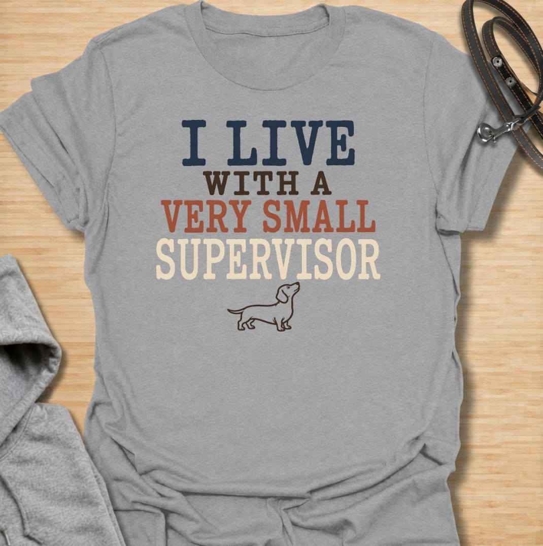 A funny dachshund T-shirt for anyone who lives with a very small dog who insists on overseeing absolutely everything. Featuring the phrase “I Live With a Very Small Supervisor” and a simple dachshund illustration, this design captures the breed’s curious, watchful personality perfectly. Ideal for dachshund moms, dachshund dads, sausage dog owners, and anyone whose little dog follows them from room to room. A playful everyday tee for fans of dachshund humor, wiener dogs, and devoted four-legged supervisors.