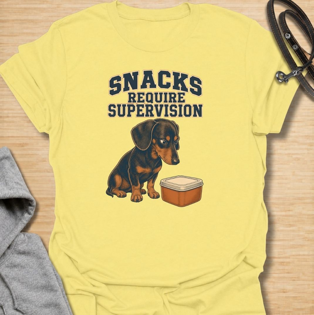 A funny dachshund T-shirt for anyone whose dog believes every snack requires close supervision. Featuring the phrase “Snacks Require Supervision” with a watchful dachshund beside a treat container, this design captures the breed’s famously food-focused personality. Perfect for dachshund moms, dachshund dads, sausage dog owners, and anyone who can’t open a snack without attracting immediate attention. A playful everyday tee for fans of dachshund humor, wiener dogs, and permanently hungry little companions.