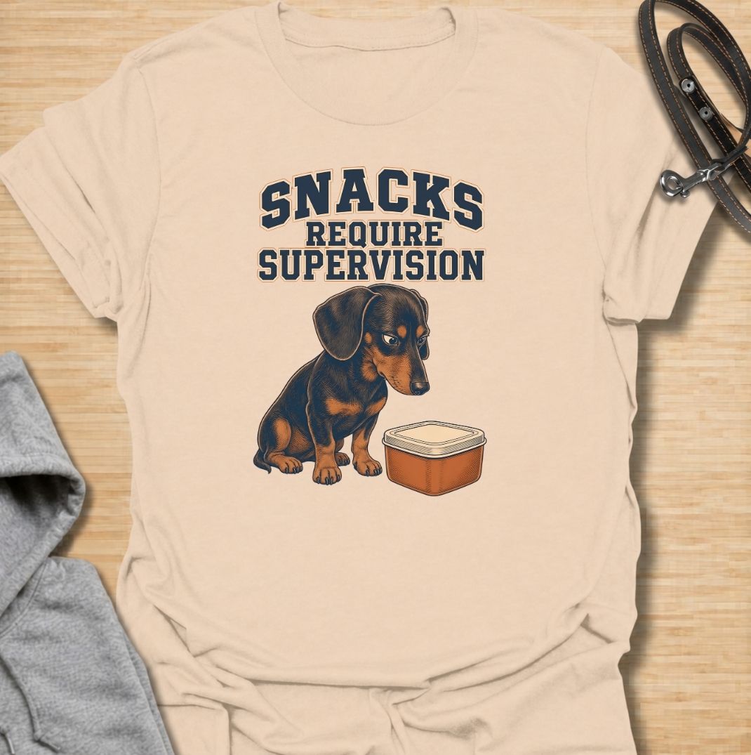 A funny dachshund T-shirt for anyone whose dog believes every snack requires close supervision. Featuring the phrase “Snacks Require Supervision” with a watchful dachshund beside a treat container, this design captures the breed’s famously food-focused personality. Perfect for dachshund moms, dachshund dads, sausage dog owners, and anyone who can’t open a snack without attracting immediate attention. A playful everyday tee for fans of dachshund humor, wiener dogs, and permanently hungry little companions.