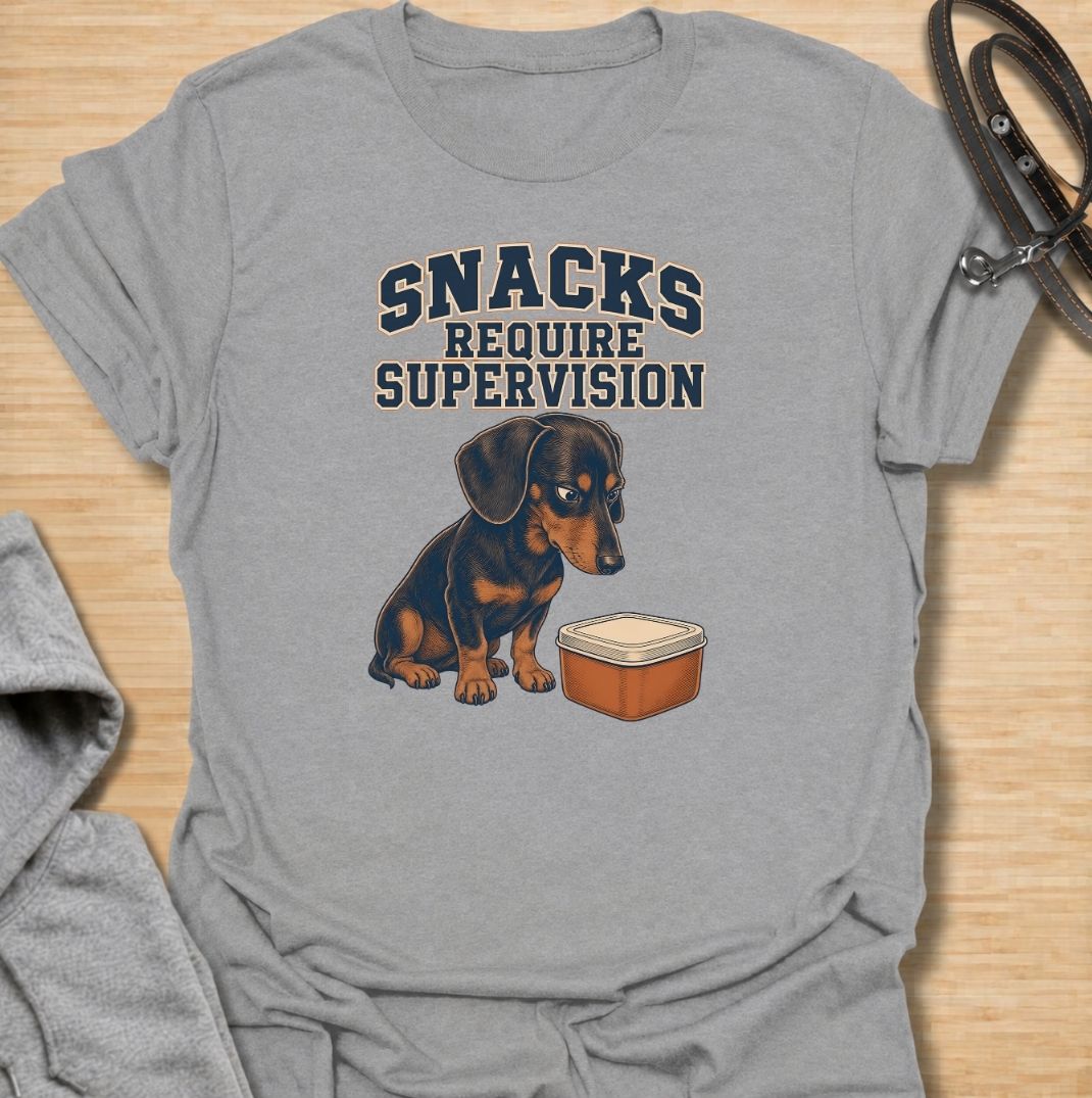 A funny dachshund T-shirt for anyone whose dog believes every snack requires close supervision. Featuring the phrase “Snacks Require Supervision” with a watchful dachshund beside a treat container, this design captures the breed’s famously food-focused personality. Perfect for dachshund moms, dachshund dads, sausage dog owners, and anyone who can’t open a snack without attracting immediate attention. A playful everyday tee for fans of dachshund humor, wiener dogs, and permanently hungry little companions.