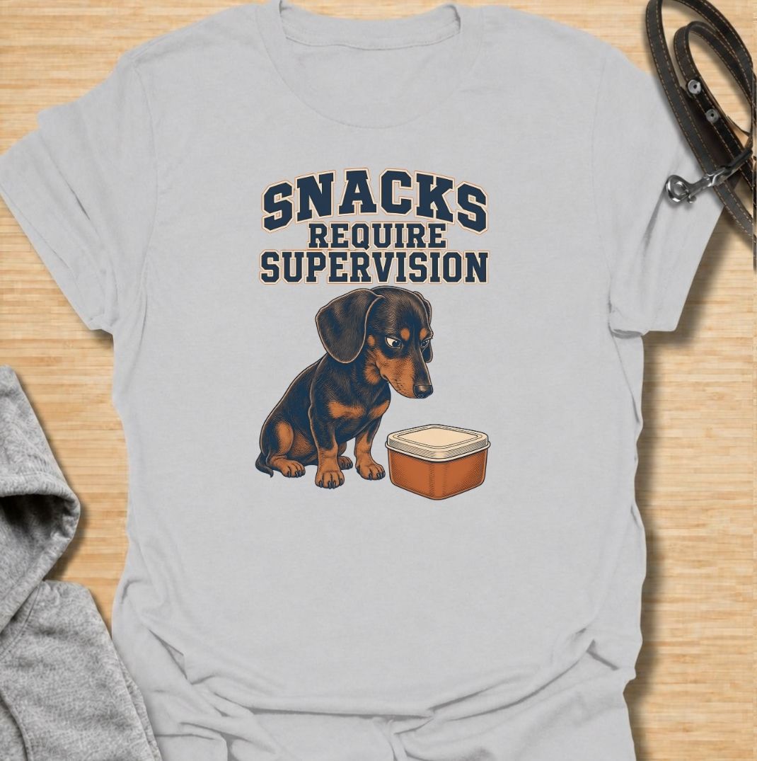 A funny dachshund T-shirt for anyone whose dog believes every snack requires close supervision. Featuring the phrase “Snacks Require Supervision” with a watchful dachshund beside a treat container, this design captures the breed’s famously food-focused personality. Perfect for dachshund moms, dachshund dads, sausage dog owners, and anyone who can’t open a snack without attracting immediate attention. A playful everyday tee for fans of dachshund humor, wiener dogs, and permanently hungry little companions.