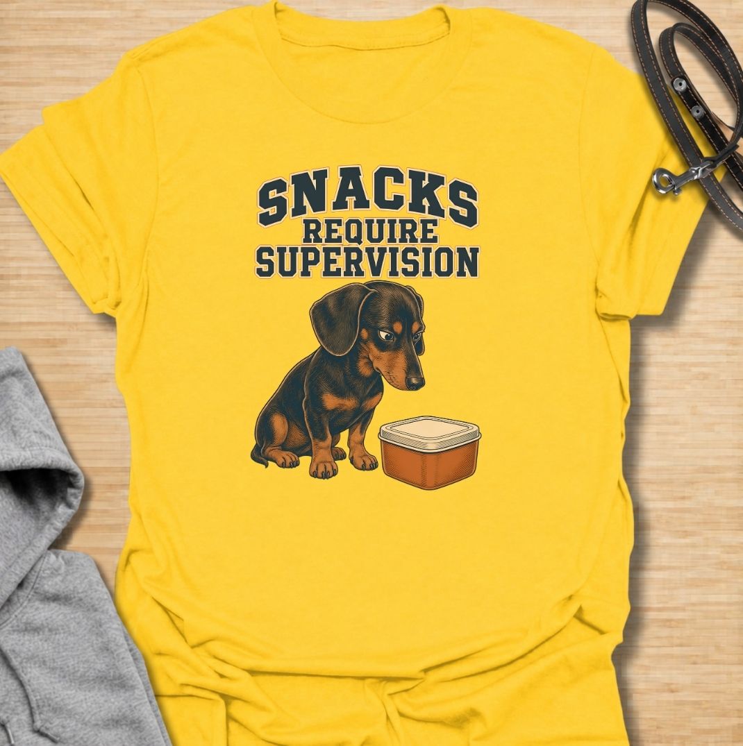 A funny dachshund T-shirt for anyone whose dog believes every snack requires close supervision. Featuring the phrase “Snacks Require Supervision” with a watchful dachshund beside a treat container, this design captures the breed’s famously food-focused personality. Perfect for dachshund moms, dachshund dads, sausage dog owners, and anyone who can’t open a snack without attracting immediate attention. A playful everyday tee for fans of dachshund humor, wiener dogs, and permanently hungry little companions.