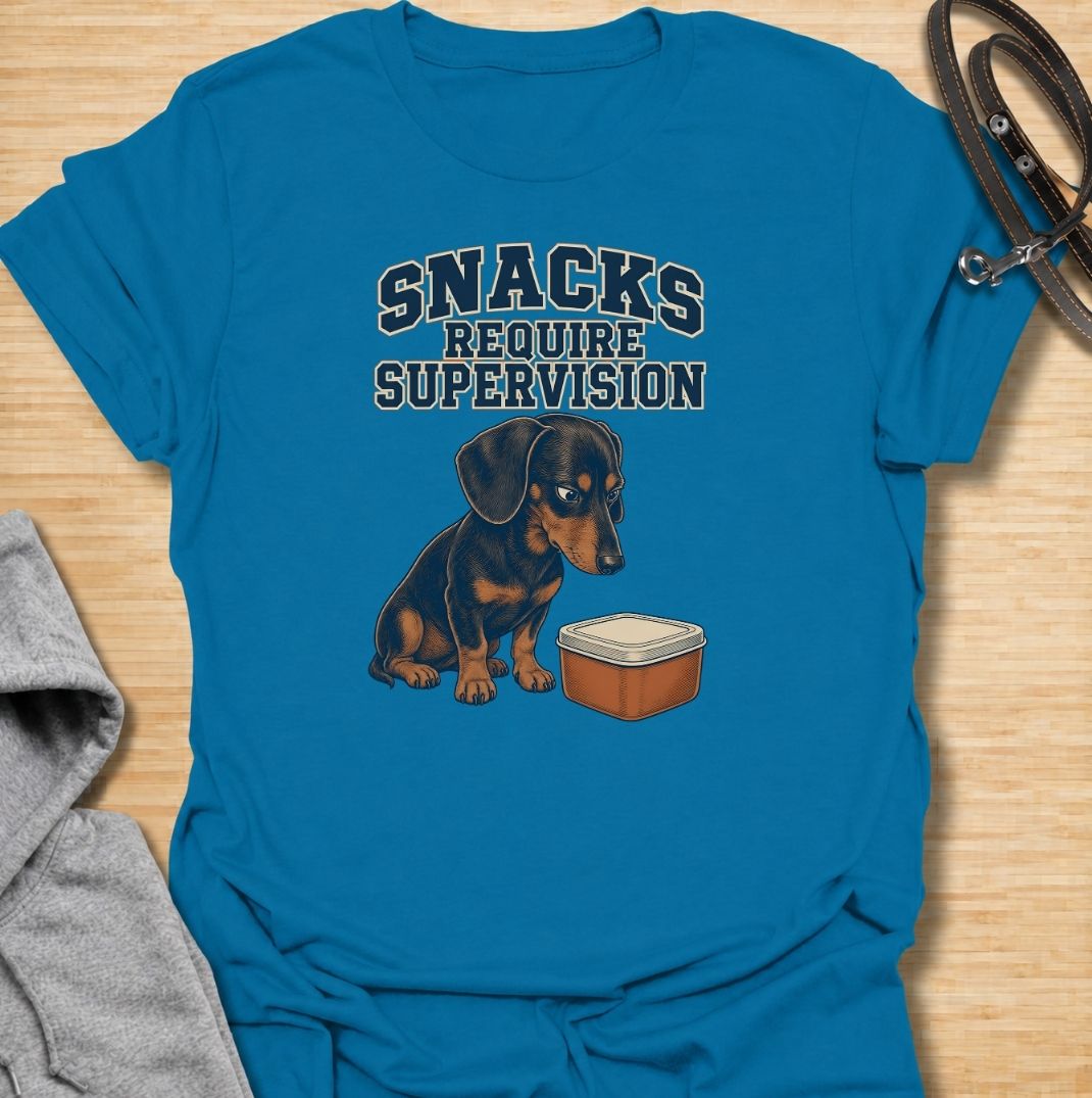 A funny dachshund T-shirt for anyone whose dog believes every snack requires close supervision. Featuring the phrase “Snacks Require Supervision” with a watchful dachshund beside a treat container, this design captures the breed’s famously food-focused personality. Perfect for dachshund moms, dachshund dads, sausage dog owners, and anyone who can’t open a snack without attracting immediate attention. A playful everyday tee for fans of dachshund humor, wiener dogs, and permanently hungry little companions.