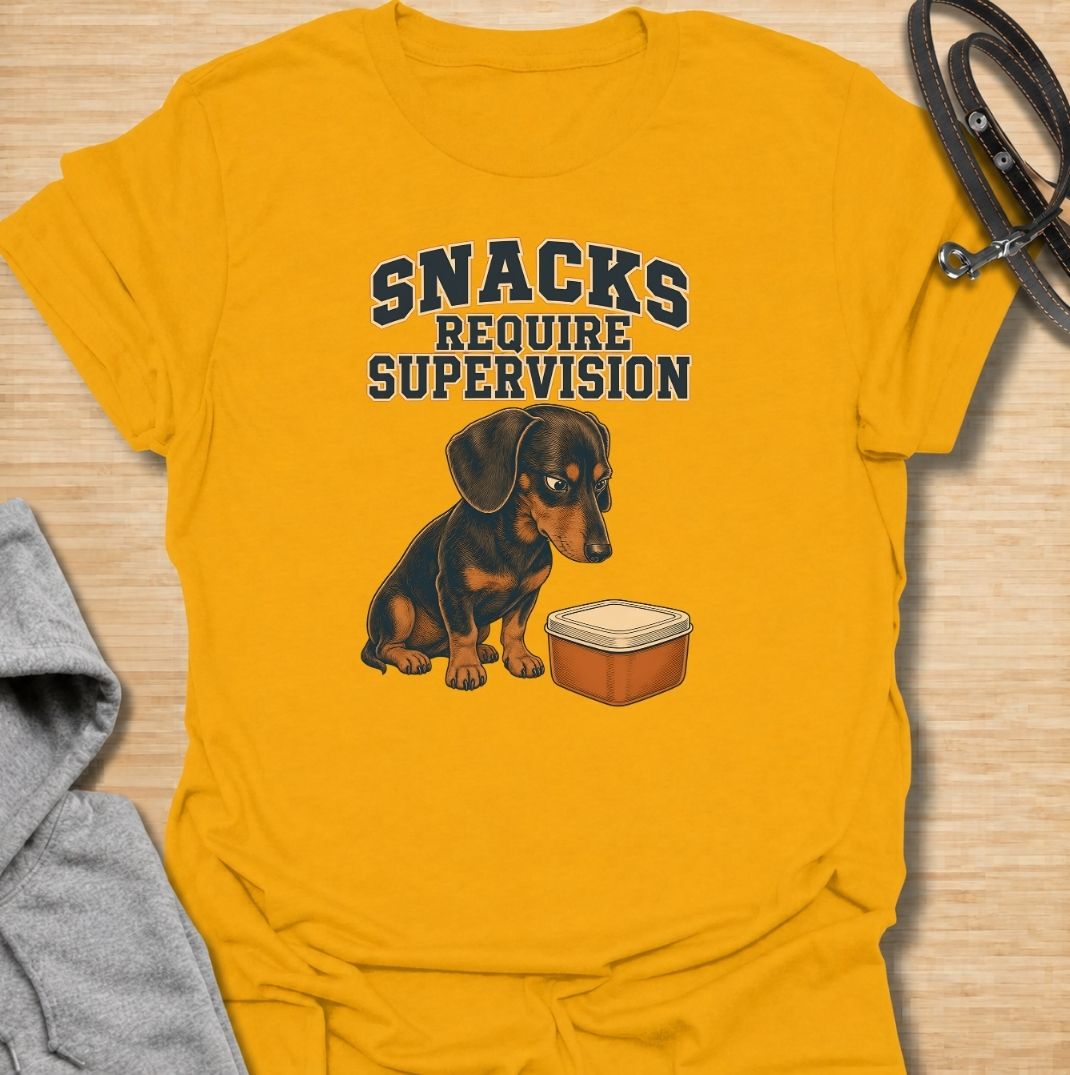 A funny dachshund T-shirt for anyone whose dog believes every snack requires close supervision. Featuring the phrase “Snacks Require Supervision” with a watchful dachshund beside a treat container, this design captures the breed’s famously food-focused personality. Perfect for dachshund moms, dachshund dads, sausage dog owners, and anyone who can’t open a snack without attracting immediate attention. A playful everyday tee for fans of dachshund humor, wiener dogs, and permanently hungry little companions.