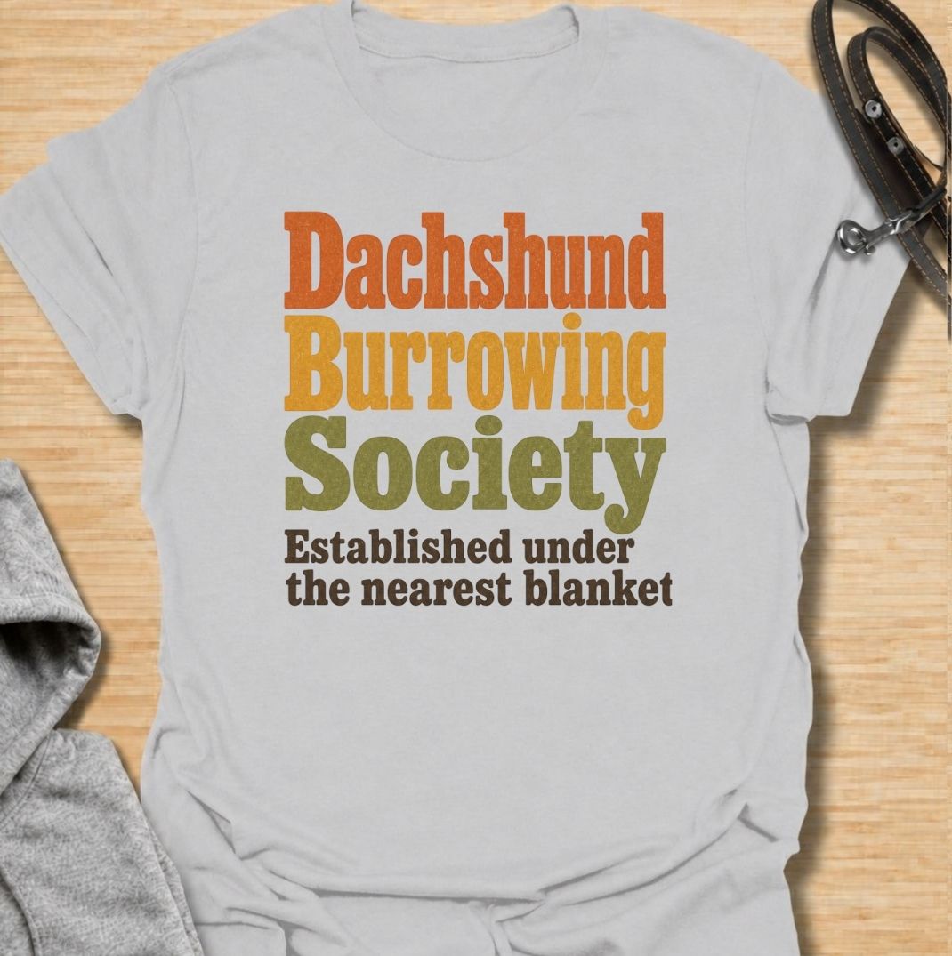 A funny dachshund T-shirt for anyone who knows the fastest way to find a sausage dog is to check under the nearest blanket. Featuring the phrase “Dachshund Burrowing Society — Established Under the Nearest Blanket,” this retro text design celebrates one of the breed’s most familiar habits. Perfect for dachshund moms, dachshund dads, doxie owners, and anyone whose dog treats blankets like a second home. A cozy, humorous everyday tee for fans of dachshund personality, burrowing behavior, and breed-specific do