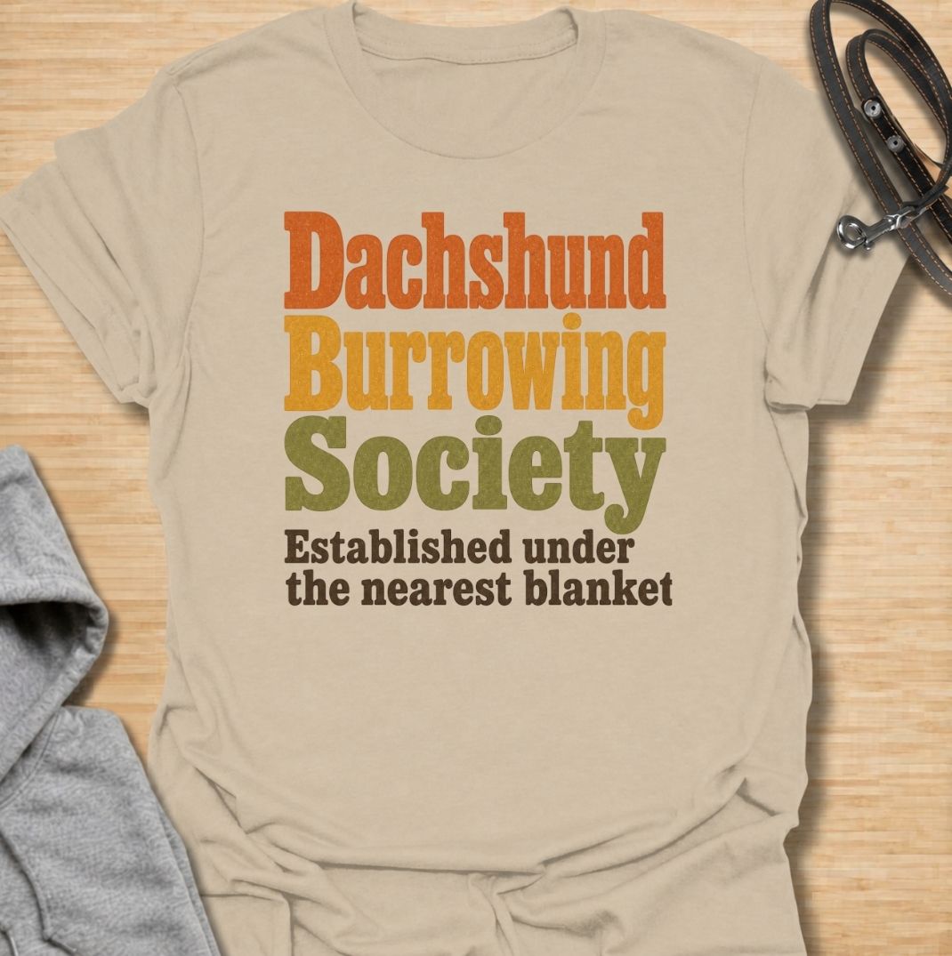 A funny dachshund T-shirt for anyone who knows the fastest way to find a sausage dog is to check under the nearest blanket. Featuring the phrase “Dachshund Burrowing Society — Established Under the Nearest Blanket,” this retro text design celebrates one of the breed’s most familiar habits. Perfect for dachshund moms, dachshund dads, doxie owners, and anyone whose dog treats blankets like a second home. A cozy, humorous everyday tee for fans of dachshund personality, burrowing behavior, and breed-specific do