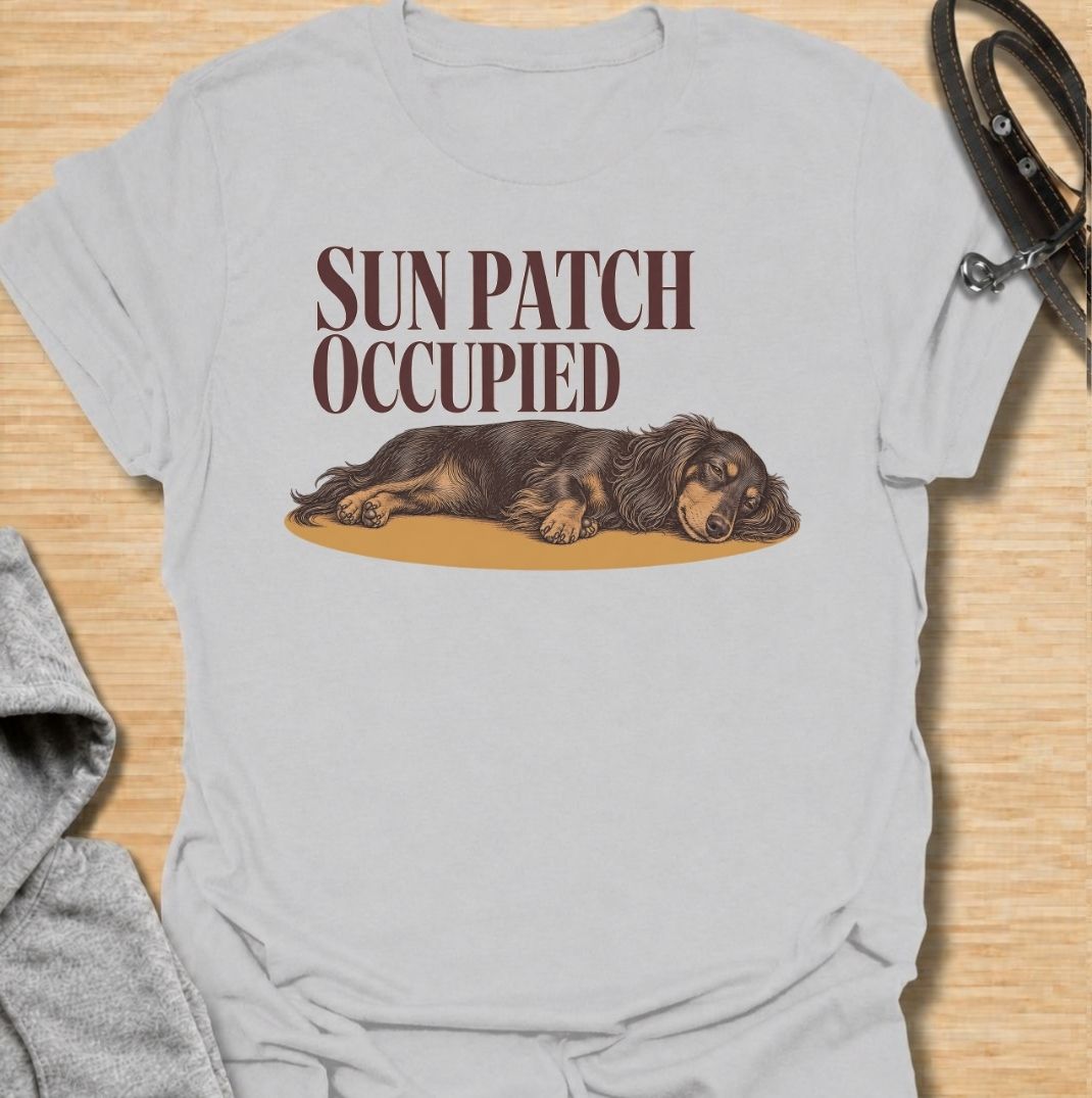 A funny dachshund T-shirt for anyone who knows the best sunny spot in the house is already taken. Featuring the phrase “Sun Patch Occupied” with a relaxed dachshund stretched out in the warmth, this design captures one of the breed’s most familiar little habits. Perfect for dachshund moms, dachshund dads, sausage dog owners, and anyone whose dog follows every patch of sunshine around the room. A cozy everyday tee for fans of dachshund humor, sleepy dogs, and breed-specific gifts.