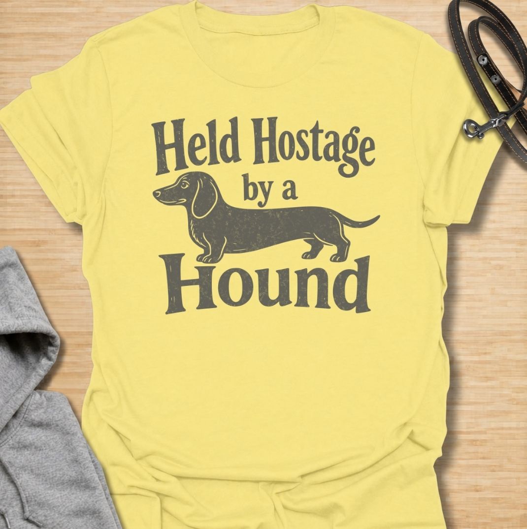 A funny dachshund T-shirt for anyone who knows these little dogs have a way of taking over your time, your seat, and your entire routine. Featuring the phrase “Held Hostage by a Hound” with a simple dachshund graphic, this design captures the breed’s clingy, demanding personality with dry humor. Perfect for dachshund moms, dachshund dads, sausage dog owners, and anyone whose dog always gets the final say. A playful everyday tee for fans of dachshund humor, doxies, and breed-specific dog gifts.