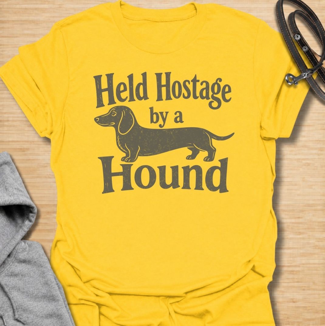 A funny dachshund T-shirt for anyone who knows these little dogs have a way of taking over your time, your seat, and your entire routine. Featuring the phrase “Held Hostage by a Hound” with a simple dachshund graphic, this design captures the breed’s clingy, demanding personality with dry humor. Perfect for dachshund moms, dachshund dads, sausage dog owners, and anyone whose dog always gets the final say. A playful everyday tee for fans of dachshund humor, doxies, and breed-specific dog gifts.