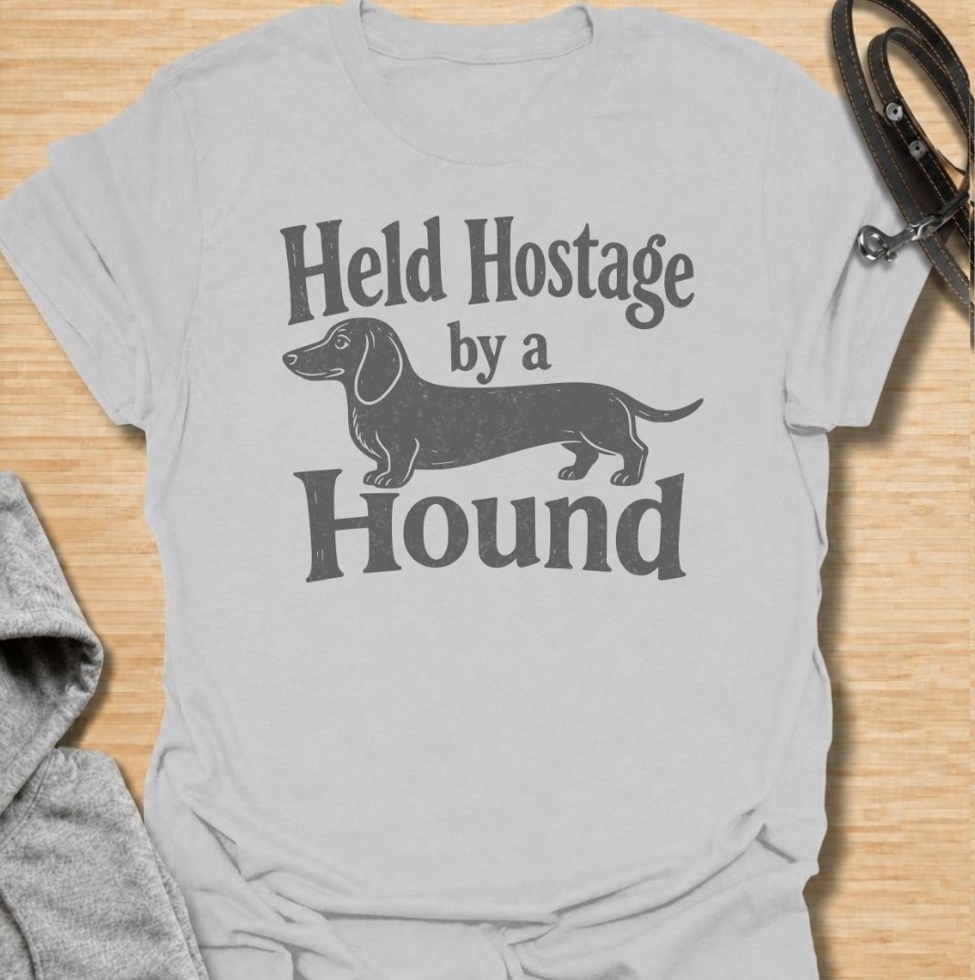 A funny dachshund T-shirt for anyone who knows these little dogs have a way of taking over your time, your seat, and your entire routine. Featuring the phrase “Held Hostage by a Hound” with a simple dachshund graphic, this design captures the breed’s clingy, demanding personality with dry humor. Perfect for dachshund moms, dachshund dads, sausage dog owners, and anyone whose dog always gets the final say. A playful everyday tee for fans of dachshund humor, doxies, and breed-specific dog gifts.