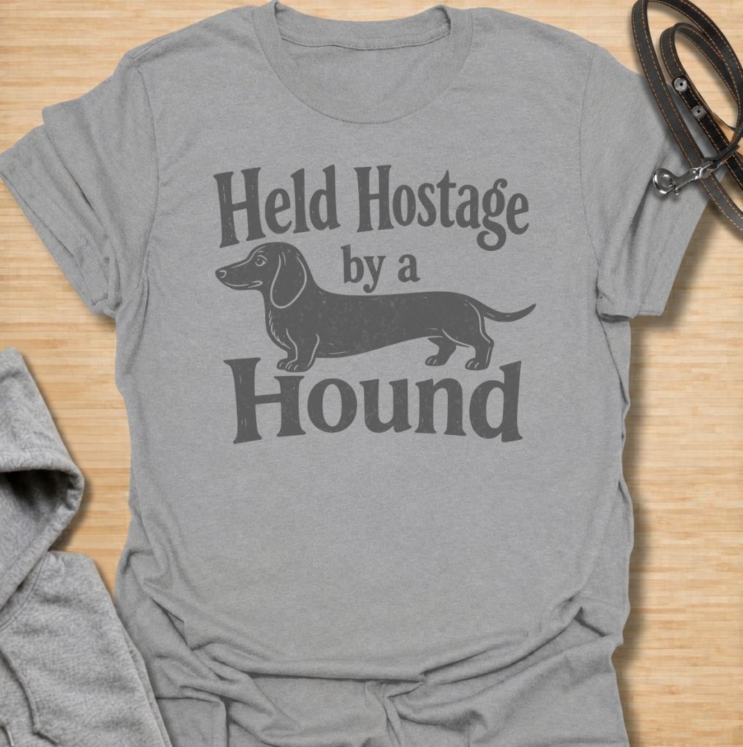 A funny dachshund T-shirt for anyone who knows these little dogs have a way of taking over your time, your seat, and your entire routine. Featuring the phrase “Held Hostage by a Hound” with a simple dachshund graphic, this design captures the breed’s clingy, demanding personality with dry humor. Perfect for dachshund moms, dachshund dads, sausage dog owners, and anyone whose dog always gets the final say. A playful everyday tee for fans of dachshund humor, doxies, and breed-specific dog gifts.