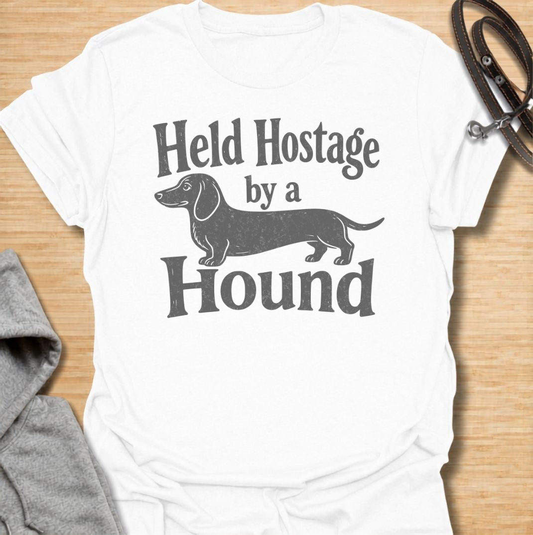 A funny dachshund T-shirt for anyone who knows these little dogs have a way of taking over your time, your seat, and your entire routine. Featuring the phrase “Held Hostage by a Hound” with a simple dachshund graphic, this design captures the breed’s clingy, demanding personality with dry humor. Perfect for dachshund moms, dachshund dads, sausage dog owners, and anyone whose dog always gets the final say. A playful everyday tee for fans of dachshund humor, doxies, and breed-specific dog gifts.