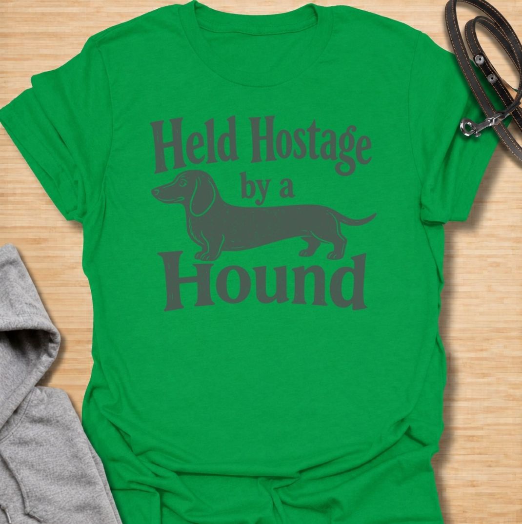 A funny dachshund T-shirt for anyone who knows these little dogs have a way of taking over your time, your seat, and your entire routine. Featuring the phrase “Held Hostage by a Hound” with a simple dachshund graphic, this design captures the breed’s clingy, demanding personality with dry humor. Perfect for dachshund moms, dachshund dads, sausage dog owners, and anyone whose dog always gets the final say. A playful everyday tee for fans of dachshund humor, doxies, and breed-specific dog gifts.