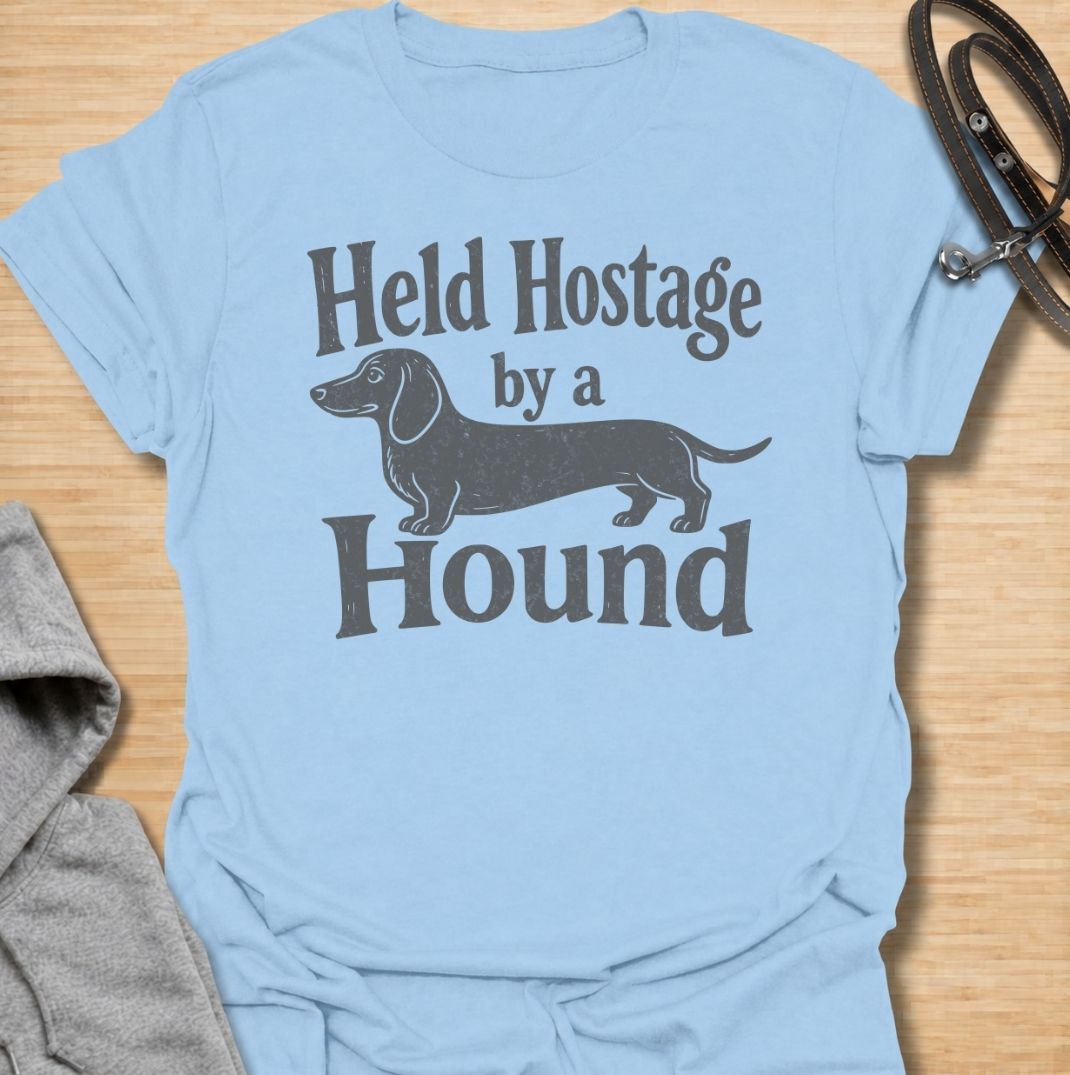 A funny dachshund T-shirt for anyone who knows these little dogs have a way of taking over your time, your seat, and your entire routine. Featuring the phrase “Held Hostage by a Hound” with a simple dachshund graphic, this design captures the breed’s clingy, demanding personality with dry humor. Perfect for dachshund moms, dachshund dads, sausage dog owners, and anyone whose dog always gets the final say. A playful everyday tee for fans of dachshund humor, doxies, and breed-specific dog gifts.