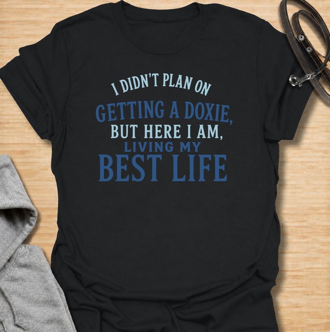 A funny dachshund T-shirt for anyone who never planned on becoming a doxie person but somehow ended up completely devoted. Featuring the phrase “I Didn’t Plan on Getting a Doxie, But Here I Am, Living My Best Life,” this text-based design captures the happy reality of dachshund ownership. Perfect for dachshund moms, dachshund dads, new doxie owners, and anyone who fell unexpectedly in love with the breed. A warm, relatable everyday tee for fans of dachshund humor, sausage dogs, and breed-specific gifts.