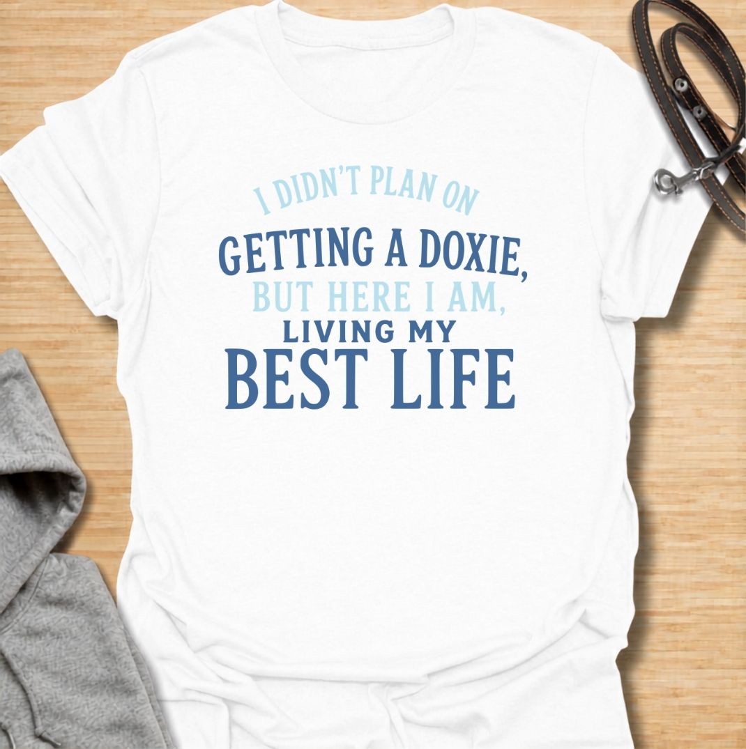 A funny dachshund T-shirt for anyone who never planned on becoming a doxie person but somehow ended up completely devoted. Featuring the phrase “I Didn’t Plan on Getting a Doxie, But Here I Am, Living My Best Life,” this text-based design captures the happy reality of dachshund ownership. Perfect for dachshund moms, dachshund dads, new doxie owners, and anyone who fell unexpectedly in love with the breed. A warm, relatable everyday tee for fans of dachshund humor, sausage dogs, and breed-specific gifts.