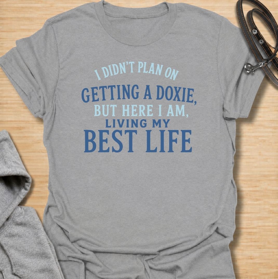 A funny dachshund T-shirt for anyone who never planned on becoming a doxie person but somehow ended up completely devoted. Featuring the phrase “I Didn’t Plan on Getting a Doxie, But Here I Am, Living My Best Life,” this text-based design captures the happy reality of dachshund ownership. Perfect for dachshund moms, dachshund dads, new doxie owners, and anyone who fell unexpectedly in love with the breed. A warm, relatable everyday tee for fans of dachshund humor, sausage dogs, and breed-specific gifts.