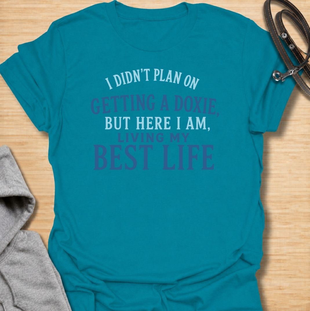 A funny dachshund T-shirt for anyone who never planned on becoming a doxie person but somehow ended up completely devoted. Featuring the phrase “I Didn’t Plan on Getting a Doxie, But Here I Am, Living My Best Life,” this text-based design captures the happy reality of dachshund ownership. Perfect for dachshund moms, dachshund dads, new doxie owners, and anyone who fell unexpectedly in love with the breed. A warm, relatable everyday tee for fans of dachshund humor, sausage dogs, and breed-specific gifts.