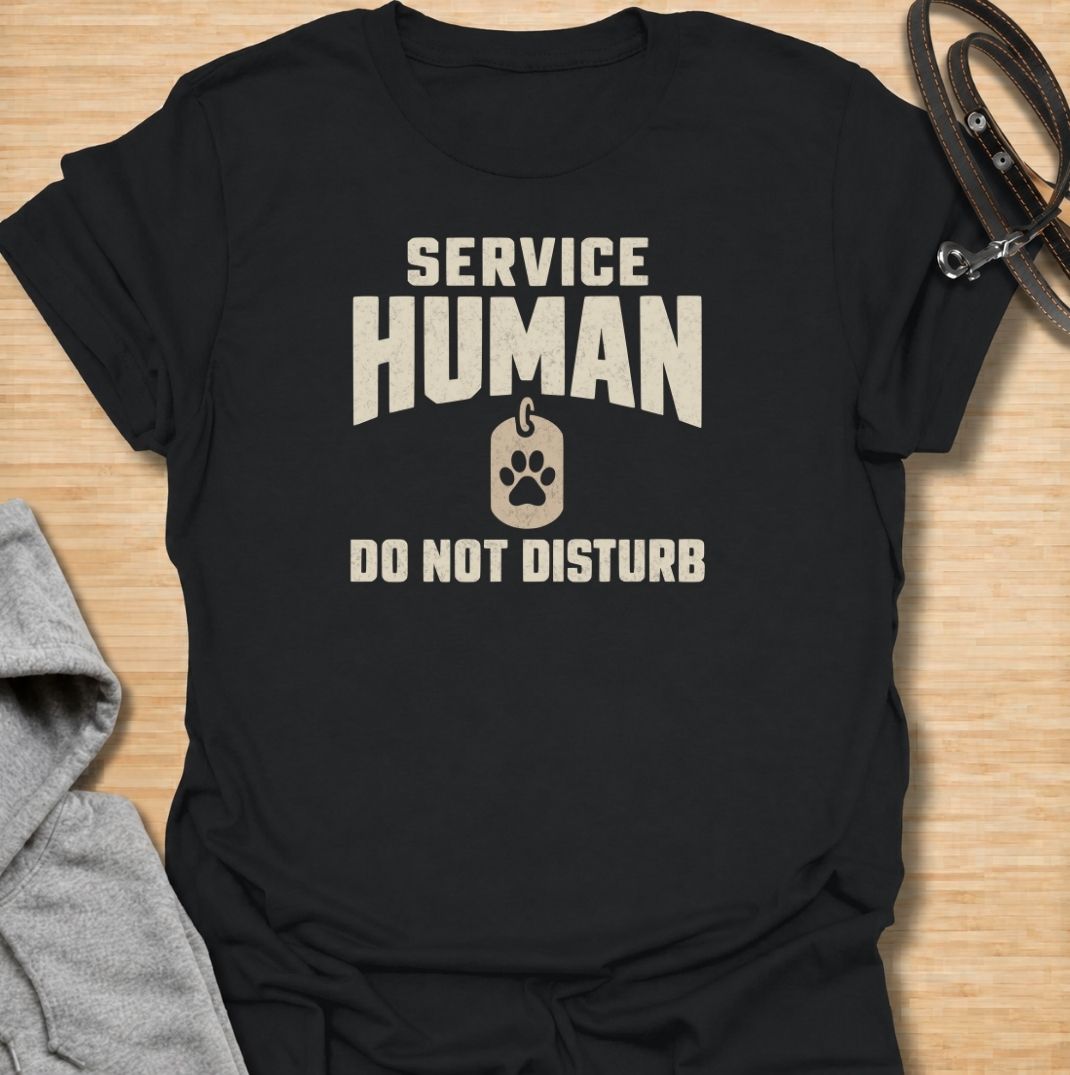 A funny dog T-shirt for anyone who knows their main role at home is serving the dog. Featuring the phrase “Service Human — Do Not Disturb” with a simple paw-print tag graphic, this clean design turns the service-dog idea on its head with dry pet-owner humor. Perfect for dog moms, dog dads, rescue dog owners, and anyone whose four-legged boss expects constant attention. A playful everyday tee for fans of funny dog sayings, pet parent humor, and general dog lover gifts.