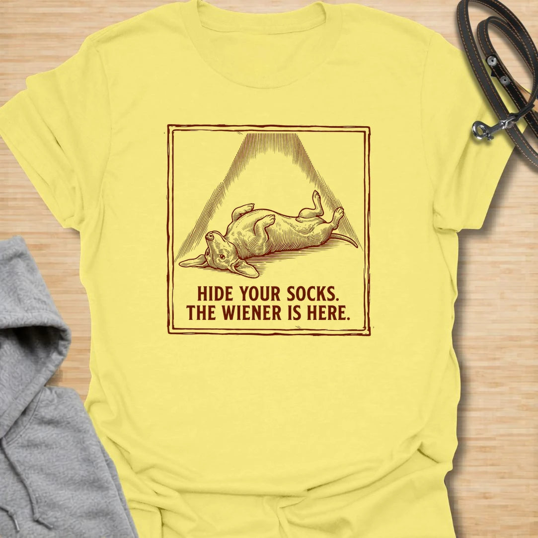 This Hide Your Socks The Wiener Is Here t-shirt is made for dachshund lovers who know their little long dog can turn laundry into a crime scene. Featuring a vintage-style dachshund under a dramatic blanket shape with bold warning text, this design has a funny mischievous dog-parent feel. Perfect for dachshund moms, dachshund dads, wiener dog fans, rescue dog lovers, and anyone whose doxie has a thing for socks.