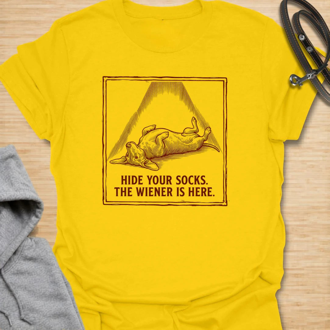 This Hide Your Socks The Wiener Is Here t-shirt is made for dachshund lovers who know their little long dog can turn laundry into a crime scene. Featuring a vintage-style dachshund under a dramatic blanket shape with bold warning text, this design has a funny mischievous dog-parent feel. Perfect for dachshund moms, dachshund dads, wiener dog fans, rescue dog lovers, and anyone whose doxie has a thing for socks.