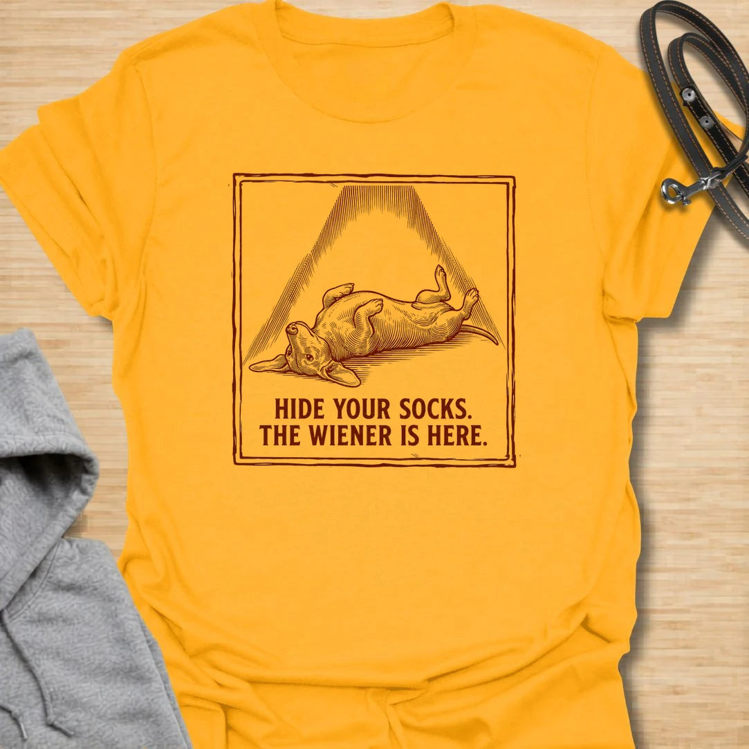 This Hide Your Socks The Wiener Is Here t-shirt is made for dachshund lovers who know their little long dog can turn laundry into a crime scene. Featuring a vintage-style dachshund under a dramatic blanket shape with bold warning text, this design has a funny mischievous dog-parent feel. Perfect for dachshund moms, dachshund dads, wiener dog fans, rescue dog lovers, and anyone whose doxie has a thing for socks.