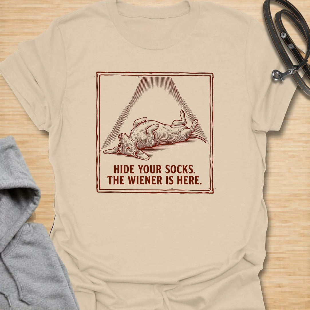 This Hide Your Socks The Wiener Is Here t-shirt is made for dachshund lovers who know their little long dog can turn laundry into a crime scene. Featuring a vintage-style dachshund under a dramatic blanket shape with bold warning text, this design has a funny mischievous dog-parent feel. Perfect for dachshund moms, dachshund dads, wiener dog fans, rescue dog lovers, and anyone whose doxie has a thing for socks.