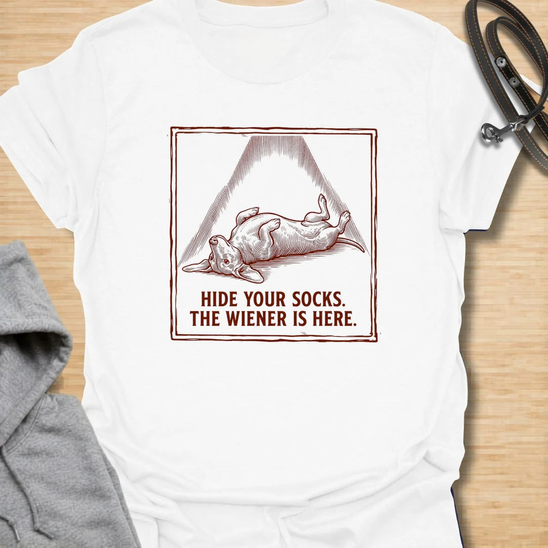 This Hide Your Socks The Wiener Is Here t-shirt is made for dachshund lovers who know their little long dog can turn laundry into a crime scene. Featuring a vintage-style dachshund under a dramatic blanket shape with bold warning text, this design has a funny mischievous dog-parent feel. Perfect for dachshund moms, dachshund dads, wiener dog fans, rescue dog lovers, and anyone whose doxie has a thing for socks.