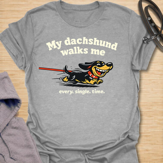 This My Dachshund Walks Me t-shirt is made for dachshund lovers who know who is really in charge of every walk. Featuring a happy running doxie pulling the leash with bold playful lettering and the phrase Every. Single. Time., this design has a funny chaotic dog-parent feel that wiener dog fans will instantly understand. Perfect for dachshund moms, dachshund dads, doxie owners, rescue dog lovers, and anyone whose tiny-legged dog sets the pace.