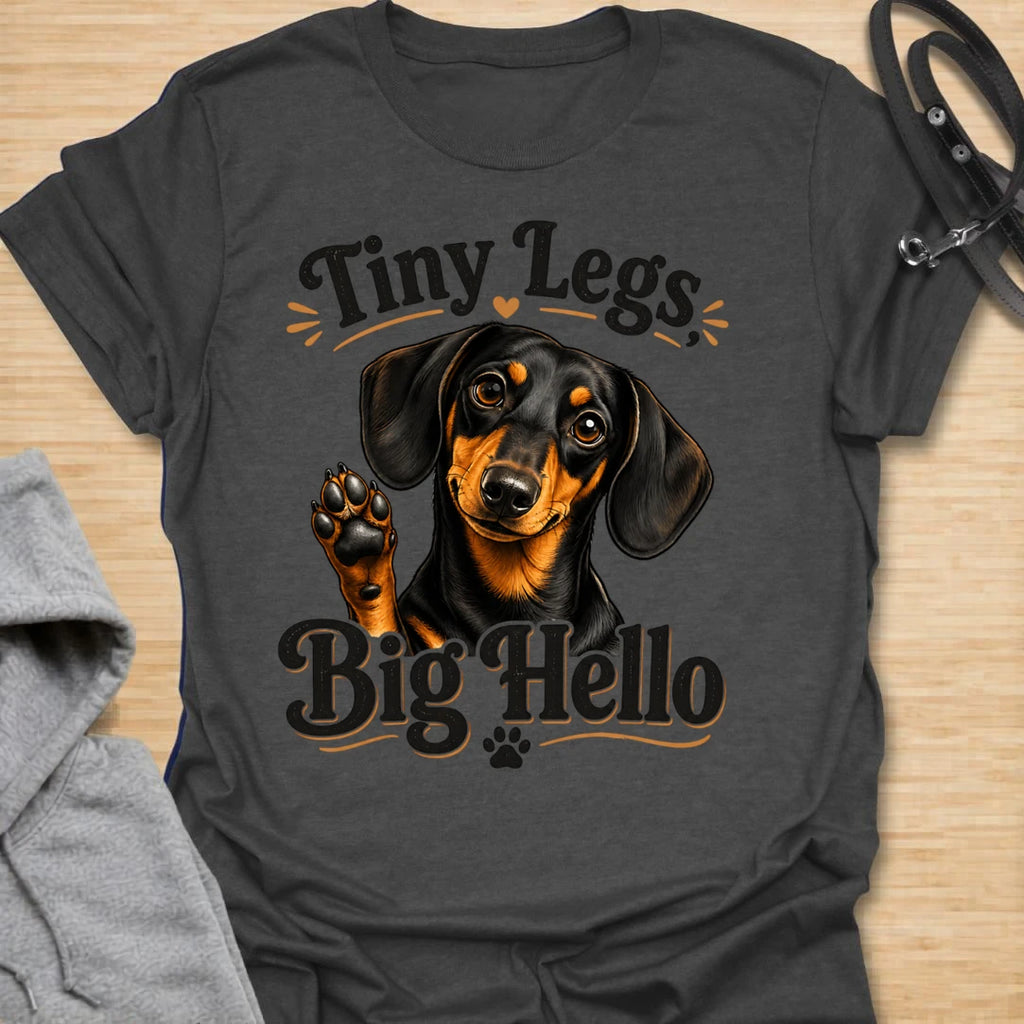 This Tiny Legs Big Hello t-shirt is made for dachshund lovers who know their little long dog can make a very big entrance. Featuring a cute black and tan dachshund waving a paw, playful retro lettering, and small heart and paw details, this design has a friendly, funny dog-lover feel. Perfect for dachshund moms, dachshund dads, wiener dog fans, doxie owners, sausage dog lovers, and anyone whose tiny-legged best friend is always ready to say hello.