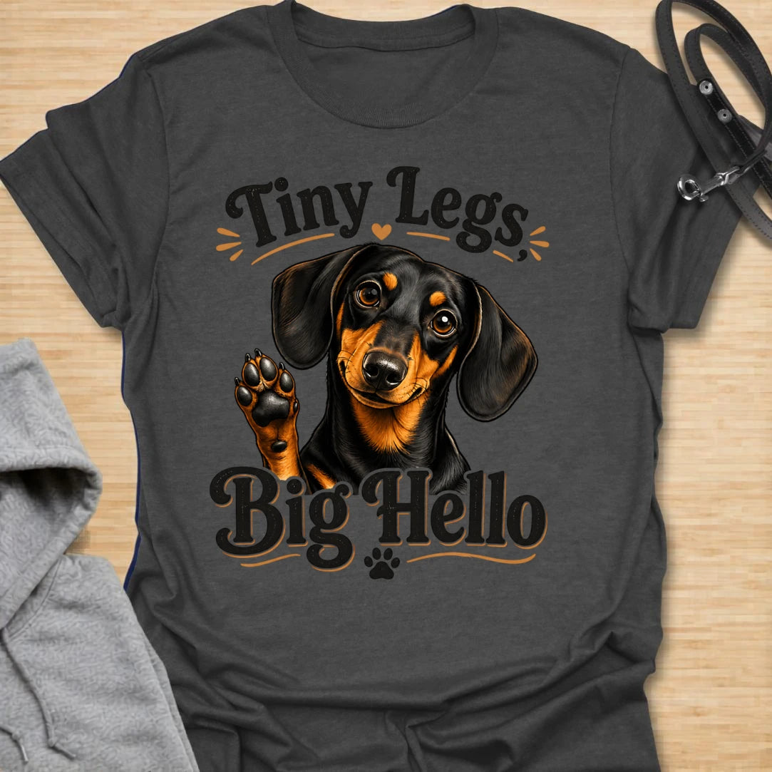 This Tiny Legs Big Hello t-shirt is made for dachshund lovers who know their little long dog can make a very big entrance. Featuring a cute black and tan dachshund waving a paw, playful retro lettering, and small heart and paw details, this design has a friendly, funny dog-lover feel. Perfect for dachshund moms, dachshund dads, wiener dog fans, doxie owners, sausage dog lovers, and anyone whose tiny-legged best friend is always ready to say hello.