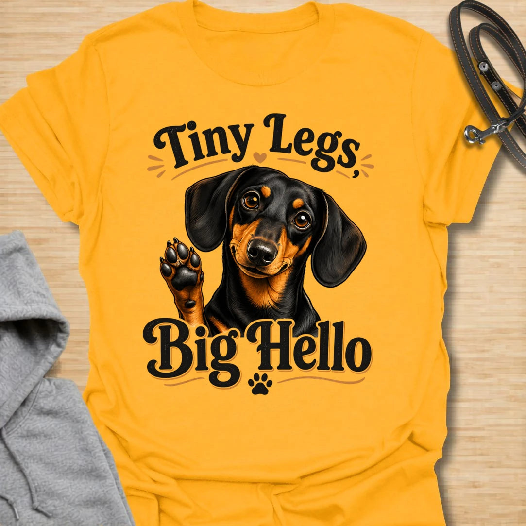 This Tiny Legs Big Hello t-shirt is made for dachshund lovers who know their little long dog can make a very big entrance. Featuring a cute black and tan dachshund waving a paw, playful retro lettering, and small heart and paw details, this design has a friendly, funny dog-lover feel. Perfect for dachshund moms, dachshund dads, wiener dog fans, doxie owners, sausage dog lovers, and anyone whose tiny-legged best friend is always ready to say hello.  Gold