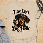 This Tiny Legs Big Hello t-shirt is made for dachshund lovers who know their little long dog can make a very big entrance. Featuring a cute black and tan dachshund waving a paw, playful retro lettering, and small heart and paw details, this design has a friendly, funny dog-lover feel. Perfect for dachshund moms, dachshund dads, wiener dog fans, doxie owners, sausage dog lovers, and anyone whose tiny-legged best friend is always ready to say hello. Sand